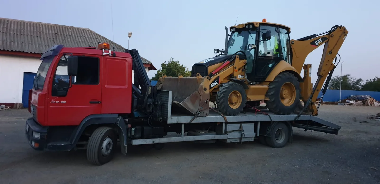 Camion MAN LE 12.180 roșu, cu macara, încărcat cu un buldoexcavator CAT 428E ancorat în chingi